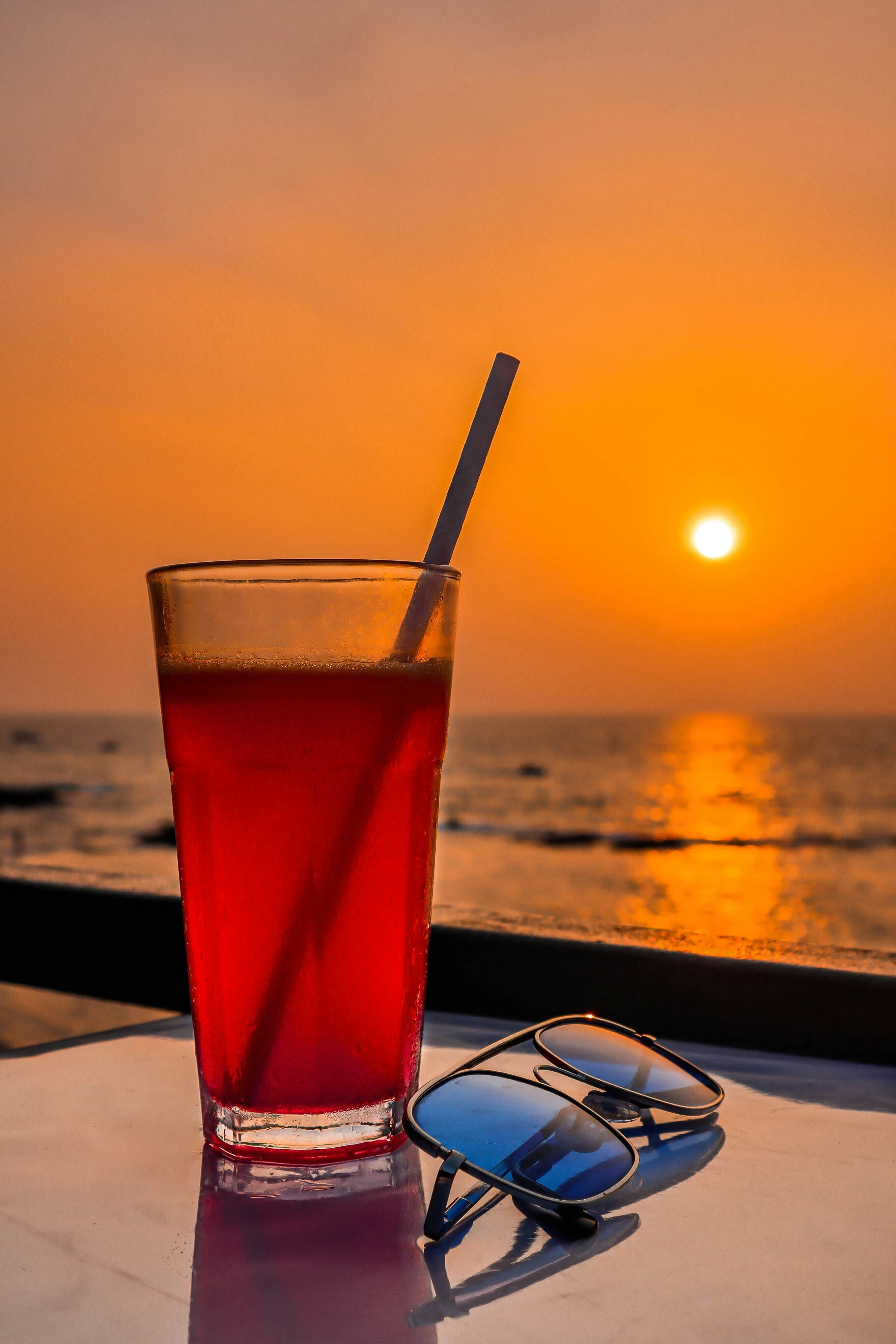 A tall red drink and sunglasses on a table as the sun sets over the sea